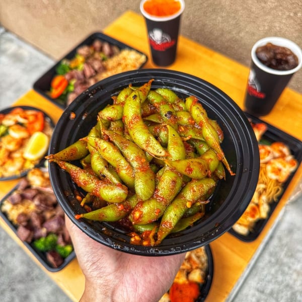 A hand holds a black bowl of spicy edamame, with other takeout containers and drinks on a wooden table.