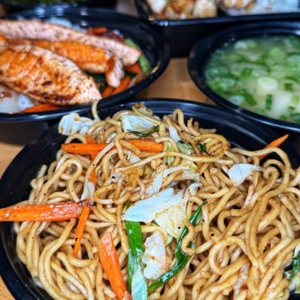 A close-up of yakisoba noodles with vegetables, salmon, and miso soup in black bowls.