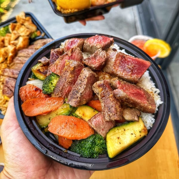 A hand holds a black bowl filled with steak, rice, carrots, zucchini, and broccoli.