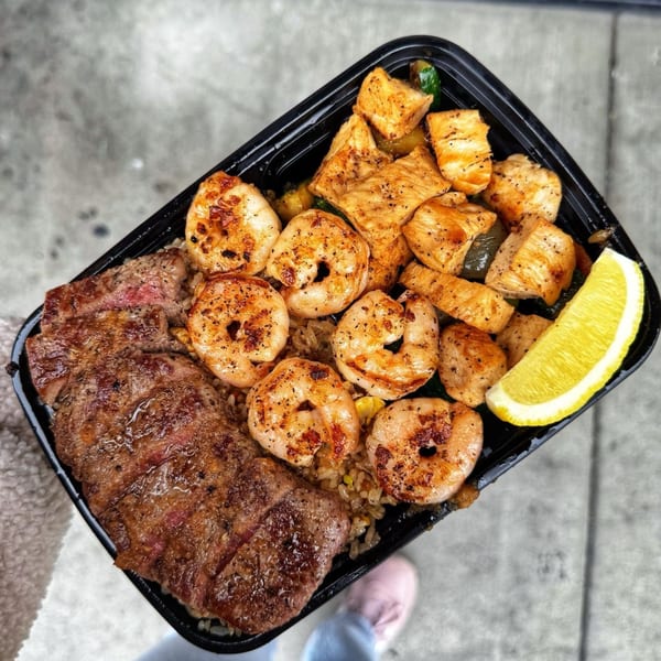 A takeout container filled with grilled steak, shrimp, chicken, and a lemon wedge.